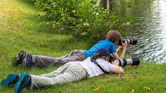 Das Bild zeigt zwei Jugendliche die am Ufer eines Sees mit einer Kamera am Boden liegen